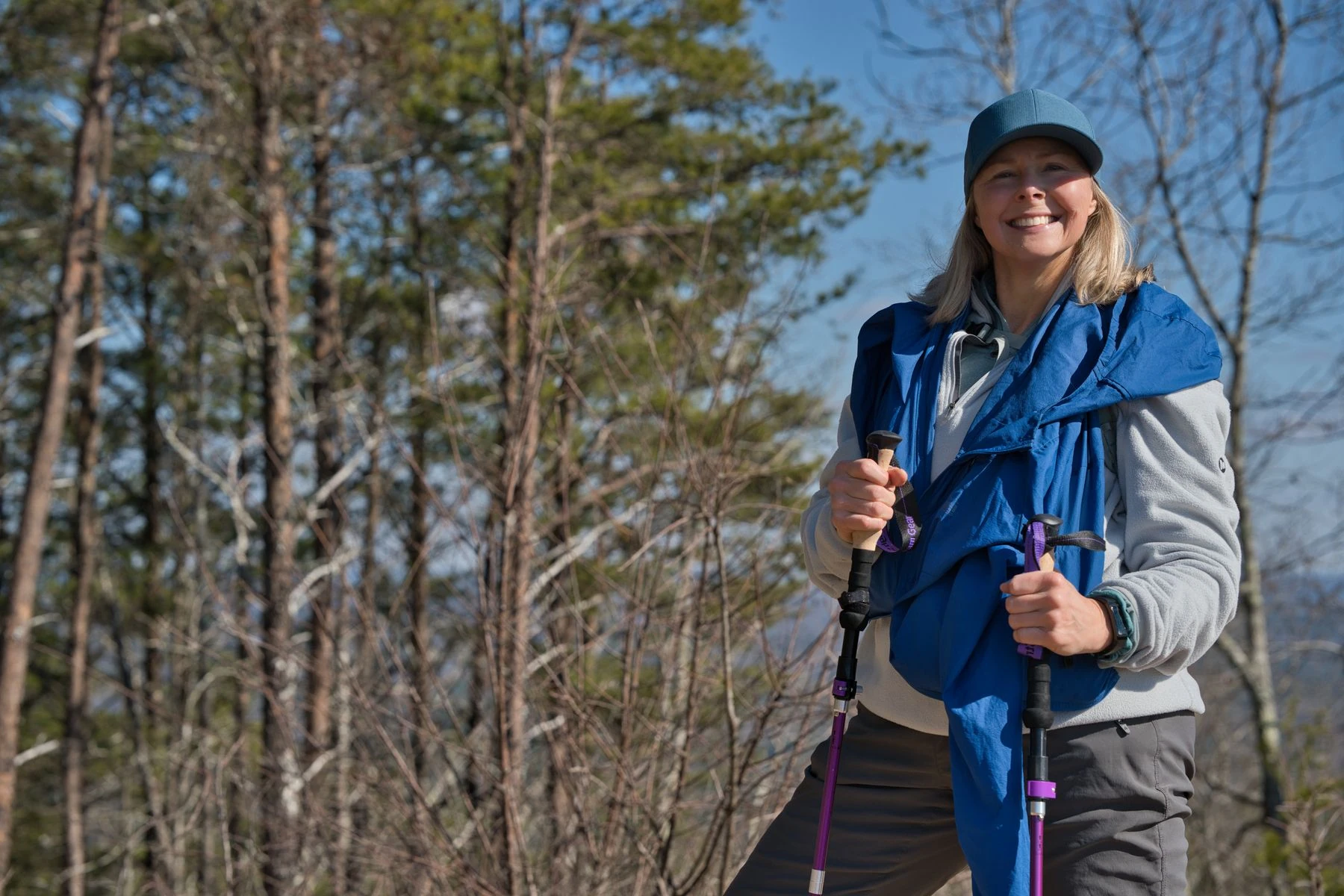 A smiling hiker holding two trekking poles on a sunny woodland trail